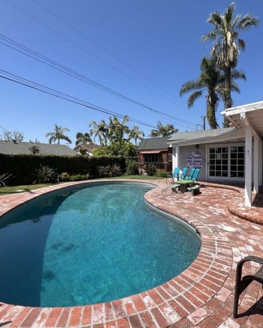 Backyard with kidney-shaped swimming pool, brick patio, white pool house, palm trees, and lounge chairs
