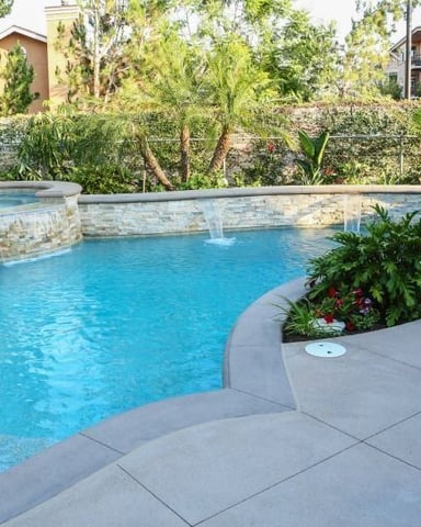 Curved swimming pool with stone waterfall and lounge chairs on concrete patio surrounded by landscaping