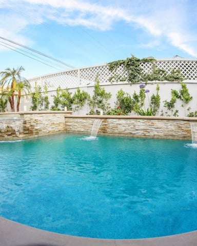 Curved swimming pool with stone waterfall feature, white wall with climbing vines, lattice fence, and palm trees