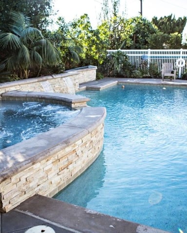 Backyard swimming pool with attached spa, stone coping, and white fence surrounded by tropical plants