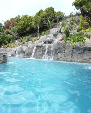 Resort pool with turquoise water, stone waterfall feature, and lush tropical landscaping on hillside