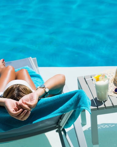 Person relaxing on lounge chair by bright blue swimming pool with drink and beach accessories nearby