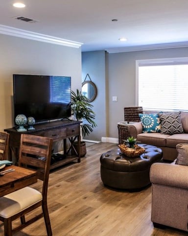 Open-plan living room with dining area, brown upholstered seating, TV on wooden stand, and large window with blinds