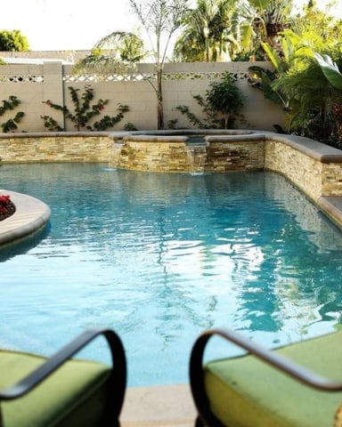 Backyard swimming pool with stone waterline, lounge chairs, tropical plants, and concrete privacy fence
