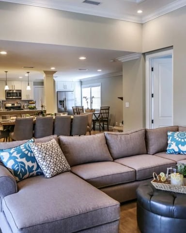 Open-plan living room with gray sectional sofa, decorative pillows, and black leather ottoman, opening to kitchen and dining area
