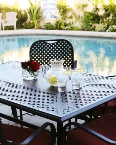 Metal patio dining table with perforated top and black chairs beside swimming pool, with potted flowers and white Adirondack chairs in background