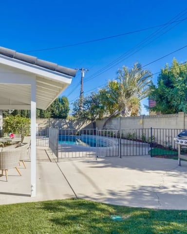 Backyard patio with covered seating area, pool with safety fence, grill, and mature landscaping under clear blue sky