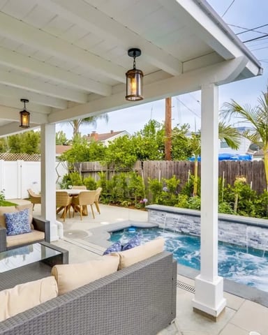 Covered patio with wicker seating, pool, and fenced backyard with palm trees and neighboring homes