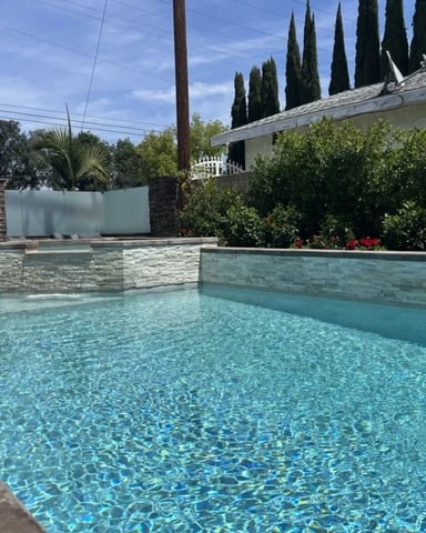 Backyard swimming pool with turquoise water, stone walls, cypress trees, and single-story house with hedges