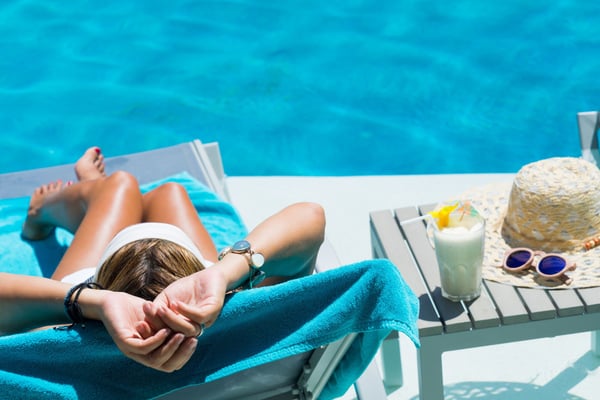Person relaxing on lounge chair by bright blue swimming pool with drink and beach accessories nearby