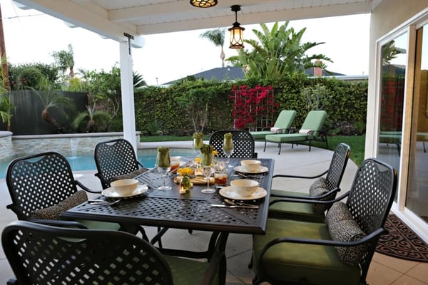 Covered patio with dining table, black chairs, pool, lounge chairs, and landscaped backyard with hedges and palm trees