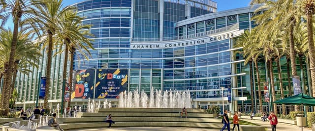 Anaheim Convention Center with modern glass facade, central fountain plaza, palm trees, and visitors on sunny day