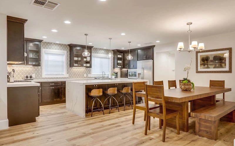 Open-plan kitchen and dining area with dark wood cabinetry, white countertops, geometric tile backsplash, island with bar seating, and wooden dining table.