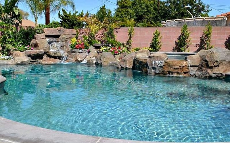 Backyard swimming pool with turquoise water, rock waterfall feature, and brick wall with landscaping