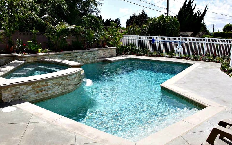 Backyard swimming pool with spa, concrete deck, stone walls, white fence, and landscaped garden