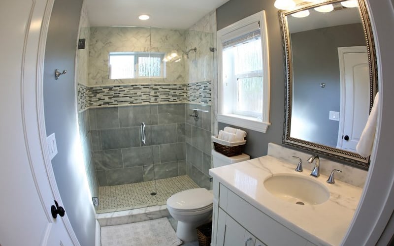 Modern bathroom with gray tile walk-in shower, white vanity with marble countertop, framed mirror, and window.