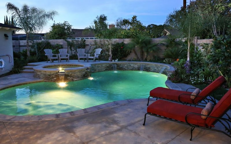 Illuminated backyard pool with spa, red lounge chairs, stone fire pit, and palm trees at dusk