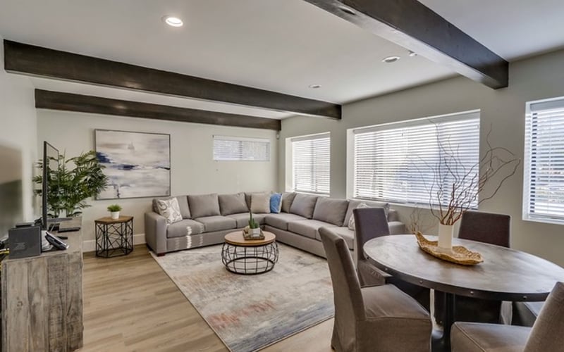 Modern living room with gray sectional sofa, round coffee table, dining area, and large windows with blinds