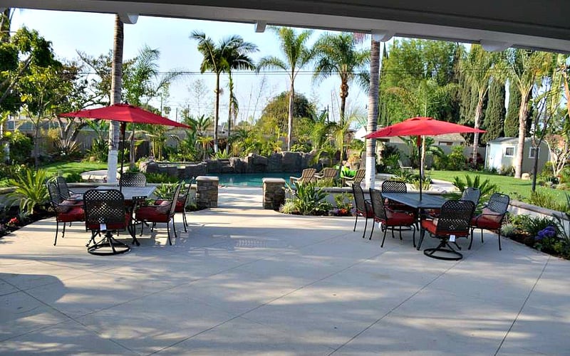 Covered patio with red umbrellas, seating areas, and pool view surrounded by palm trees and tropical landscaping