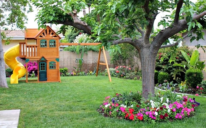 Backyard with wooden playhouse, yellow slide, swing set, large tree, and colorful flower beds