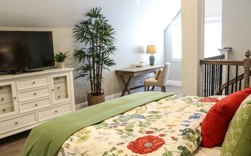 Bedroom with floral bedding, cream dresser with TV, potted plant, desk with lamp, and crib visible in background