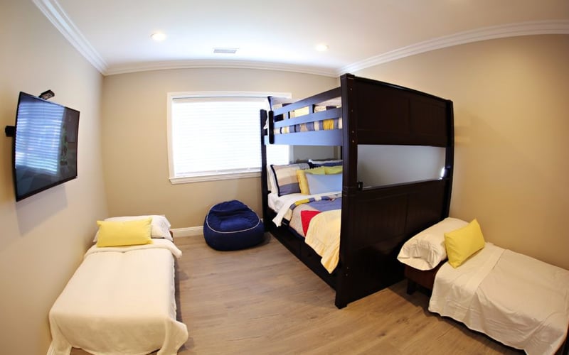 Bedroom with four beds including black bunk bed, two single beds with yellow pillows, blue bean bag, wall-mounted TV, and window with blinds.