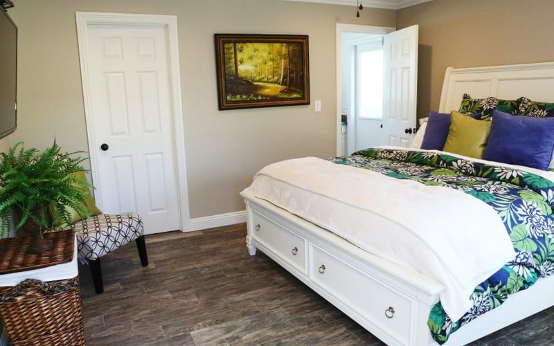 Bedroom with white bed, tropical patterned bedding, blue and yellow pillows, wood flooring, beige walls, and potted plant