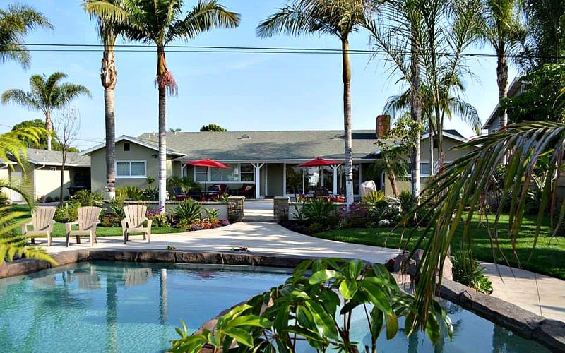 Single-story house with pool, palm trees, red umbrellas, and Adirondack chairs in tropical landscaped yard