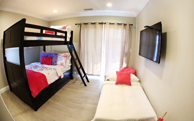 Bedroom with black bunk bed, single bed with red pillows, wall-mounted TV, and cream curtains