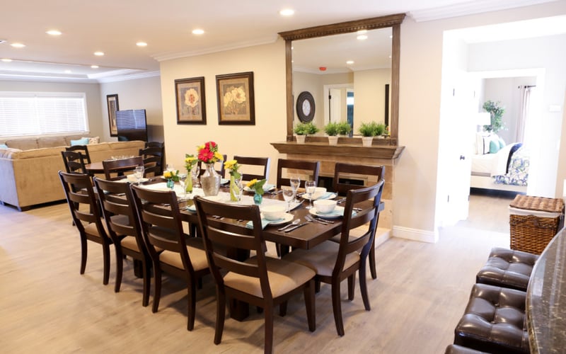 Dining room with dark wood table, six chairs, and floral centerpiece, opening to living room and kitchen beyond