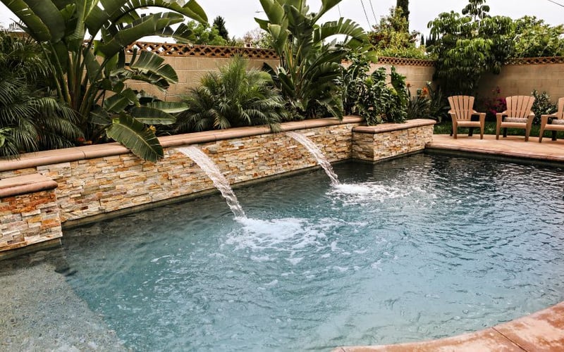 Swimming pool with stone walls and waterfall features, surrounded by tropical plants and orange lounge chairs