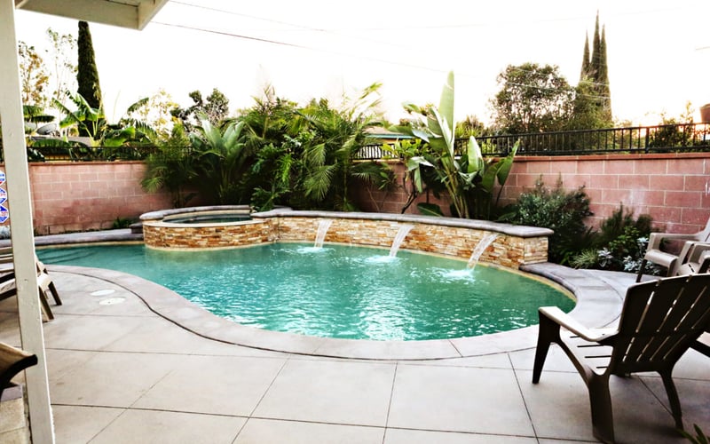 Curved swimming pool with turquoise water and stone waterfall feature, surrounded by lounge chairs and tropical plants behind brick wall